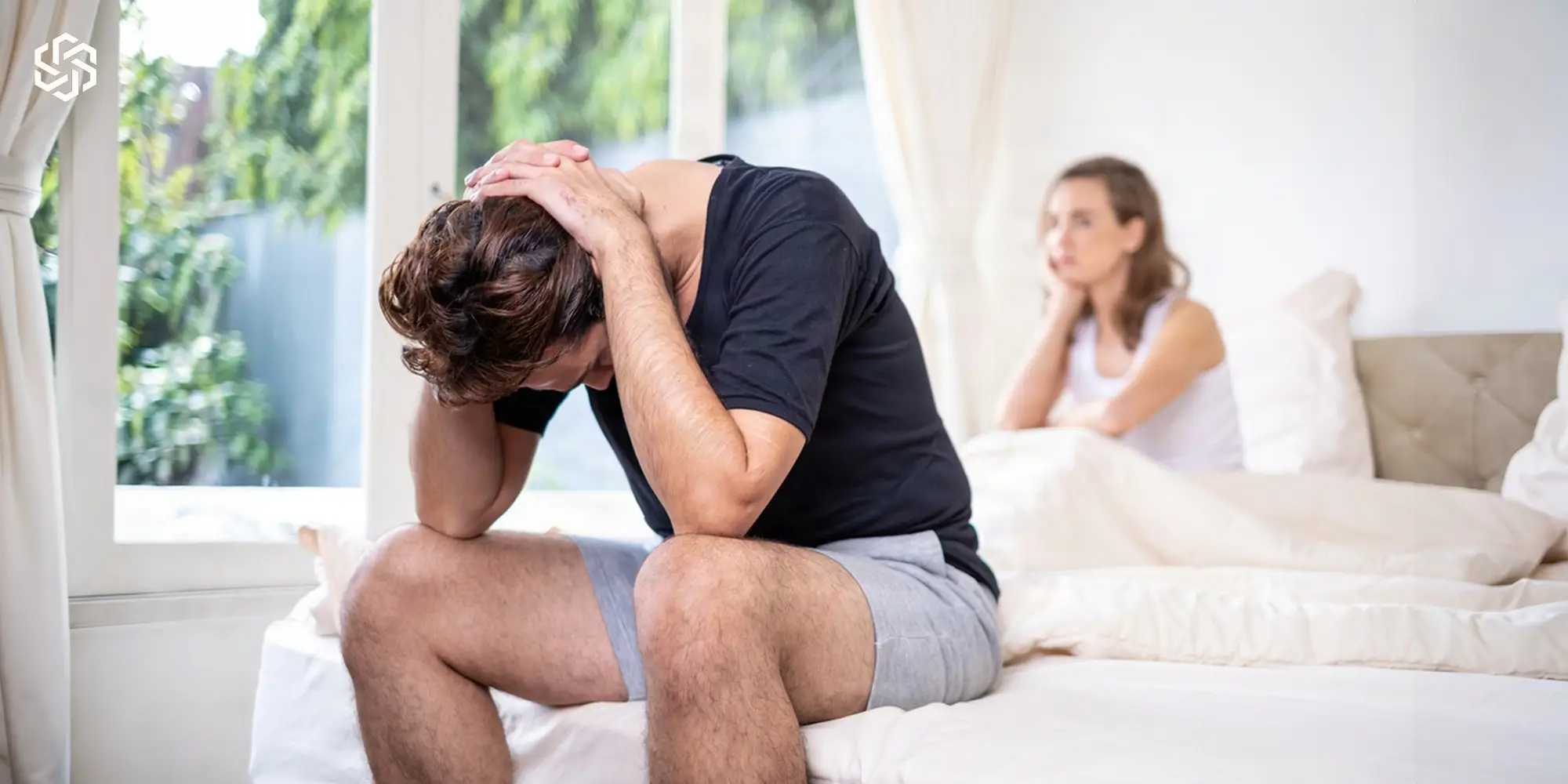 Man sitting on the edge of a bed with his head in his hands while his partner rests behind him