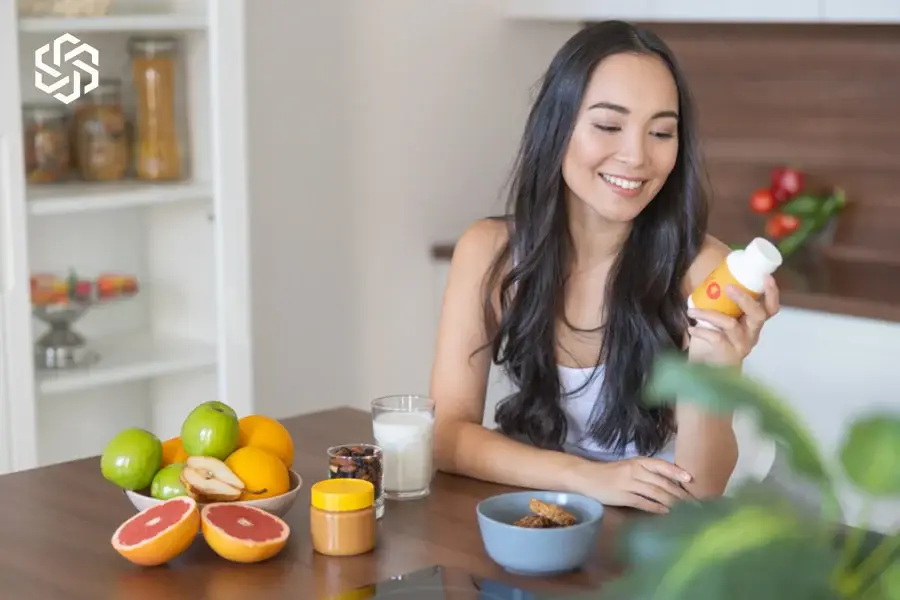 Woman holding a supplement bottle at a kitchen table with fruit