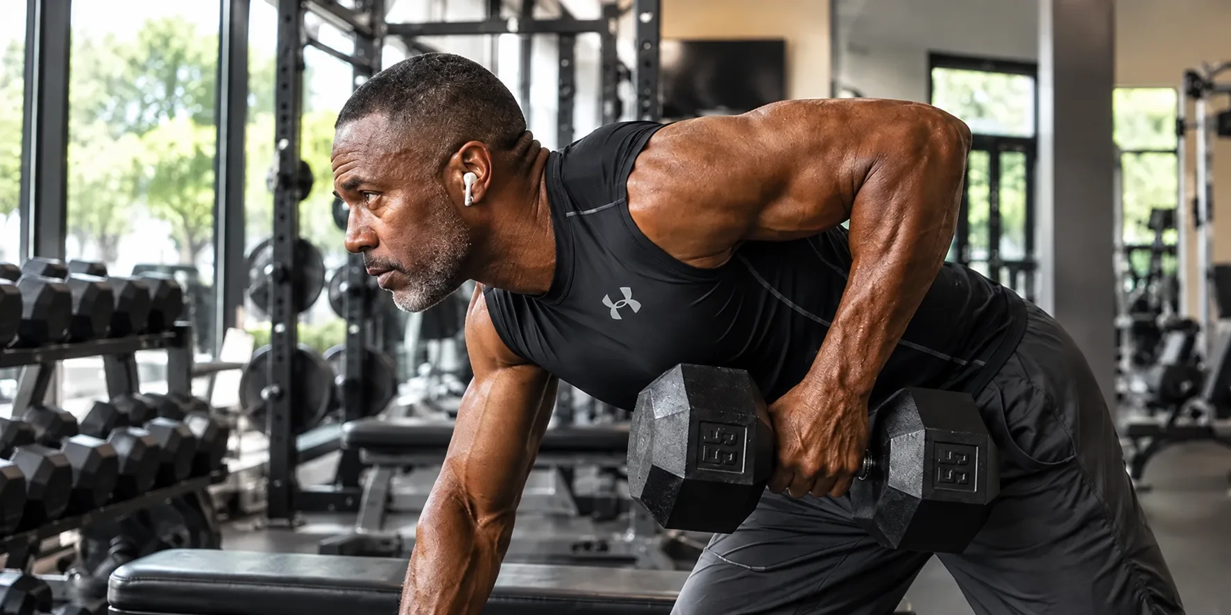 Fit man in his 50s doing a single-arm dumbbell row on a bench in a bright gym